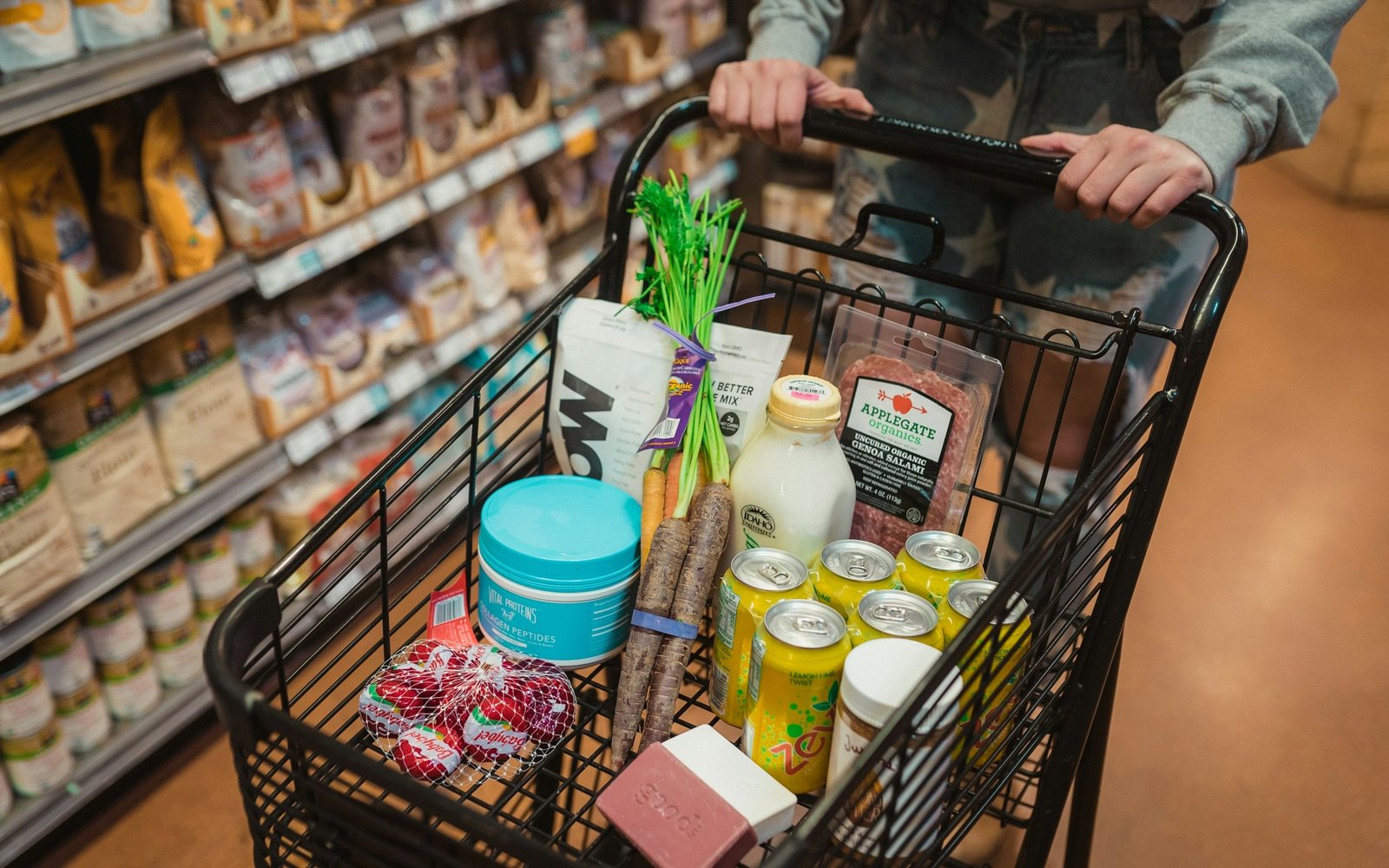 A shopper pushes a grocery cart full of food through a brightly lit store aisle.
